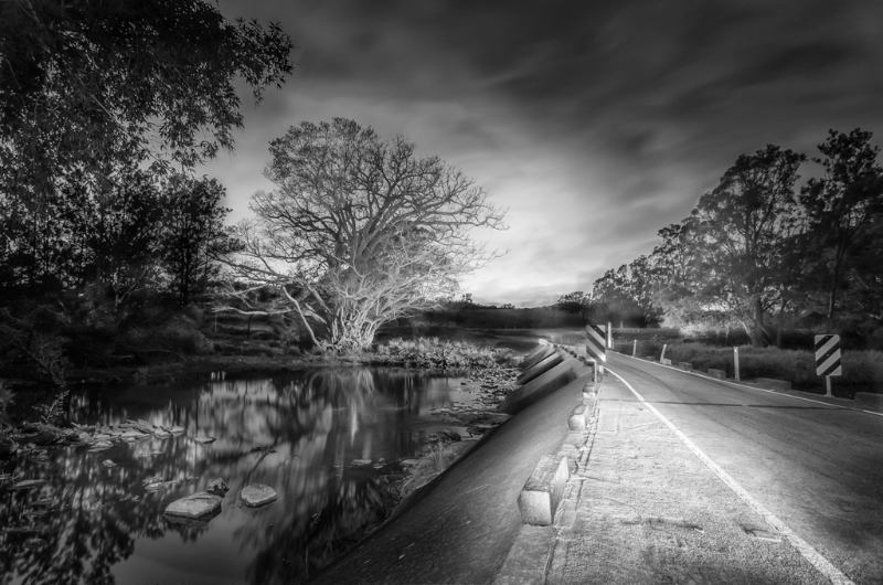 A monochrome image of a road winding and alongside a serene river, creating a captivating contrast.